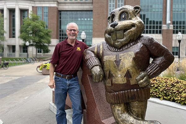 Stephen Hauser leans against a statue of goldy, both wearing  U of M maroon and gold