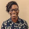 Shari Brightly-Brown wears glasses and a blue button down shirt with a tan leaf pattern. Shari is Black and has short dark brown braids combed to one side.