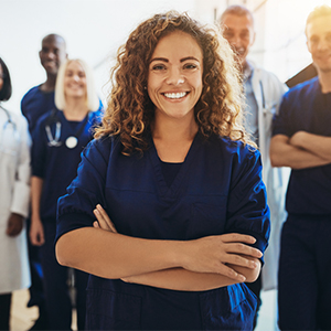 A young Latinx woman wearing a dark blue blazer stands with arms crossed with a multicultural group of health care professionals standing behind her in the background