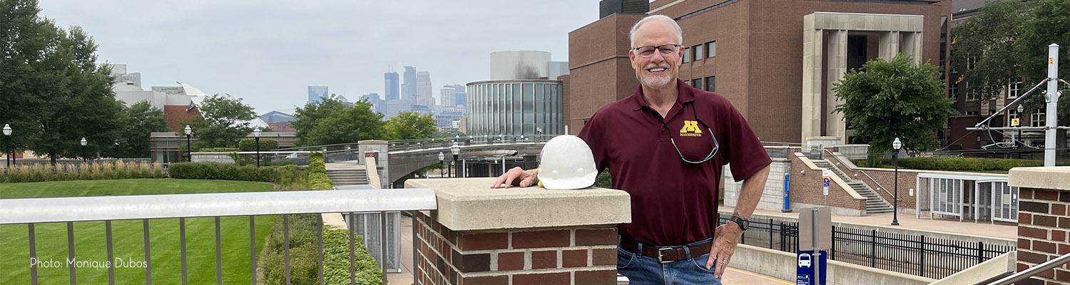 Stephen Hauser stands with his right hand on a brick newel with the East Bank campus of the U of M and the Minneapolis skyline behind him
